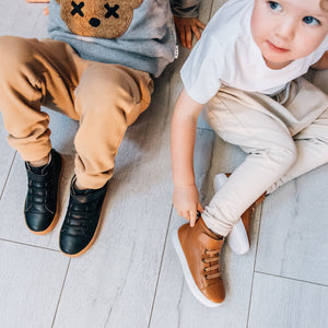 Cute boy sitting on the floor next to another child both wearing leather boots. One brown boots, one with black boots.