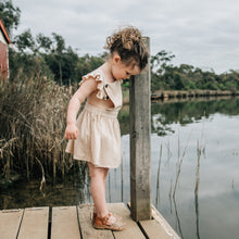 Young girl wearing beige dress and tan sandal standing by a lake.